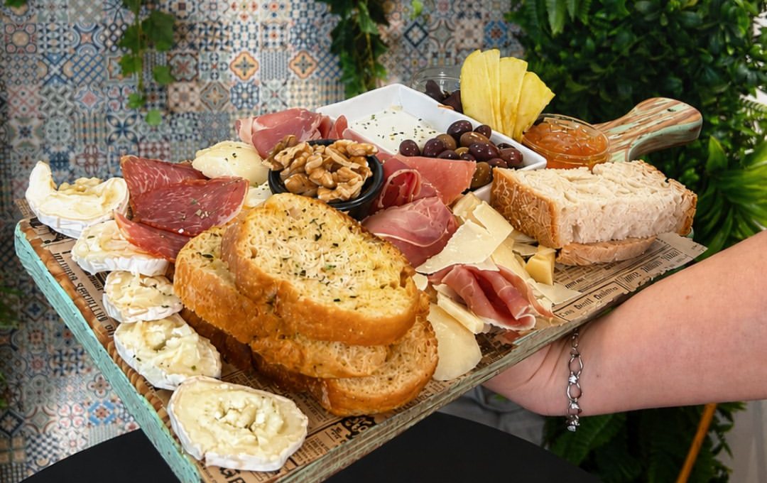 Tábua de madeira com fatias de presunto e de queijo, nozes, azeitonas, doce e pão de alho, segurada por uma pessoa